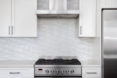 Modern bir beyaz mutfak, açık mavi ve beyaz mozaik bir arka sıçramaya karşı paslanmaz bir çelik gaz sobasını içerir.. 