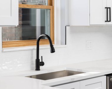 Modern bir mutfak lavabosunun yakınında siyah bir musluk, beyaz metro fayansları ve beyaz kuvars tezgahı var..