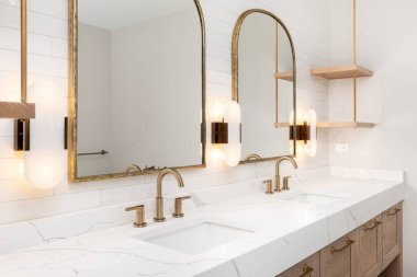 Banyoda altın musluklar, lambalar, aynalar, ahşap bir dolap, büyük mermer tezgah ve metro fayans duvarı olan bir detay..