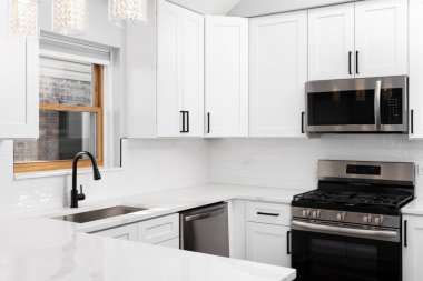 Paslanmaz çelik aletler, siyah musluk, metro döşemesi ve mermer tezgahın üzerinde asılı kolye ışıkları olan beyaz bir mutfak detayı..