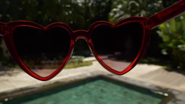 Nahaufnahme einer Sonnenbrille mit roten Herzen auf dem Hintergrund eines Swimmingpools im Luxushotel, Indonesien, Insel Bali, Zeitlupe, 4k  