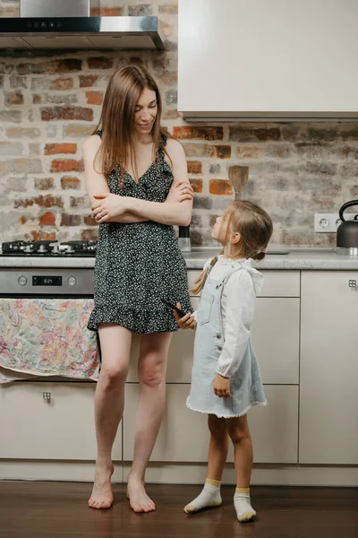 A blonde girl is keeping a smartphone while asking her mother with pale skin for something in the kitchen. A caucasian mom is standing near a pretty daughter which is using a cellphone at home.