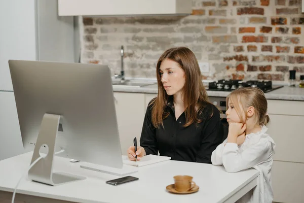 A young mother is working remotely at home while a daughter is watching it. A businesswoman is doing notes while working from an apartment near her blonde child.