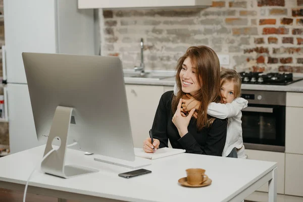 A young mother is working remotely at home while a daughter is hugging her. A smiling businesswoman is doing notes while working from an apartment near her blonde child.