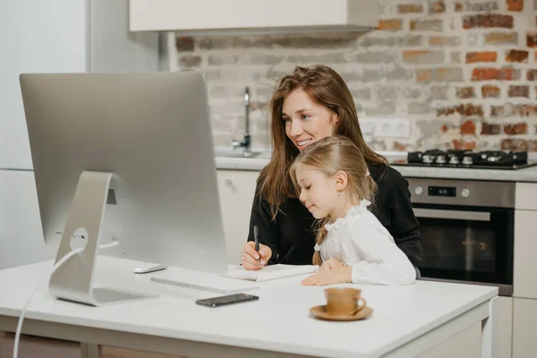 A young mother is working remotely at home while a daughter is studying from her. A smiling businesswoman is taking notes while working from an apartment near her blonde child.