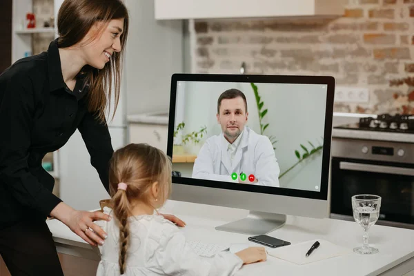 A young mother and daughter during an online doctor\'s appointment via a video call on the desktop computer. A mom and a blonde child get a doctor\'s remote consultation at home.