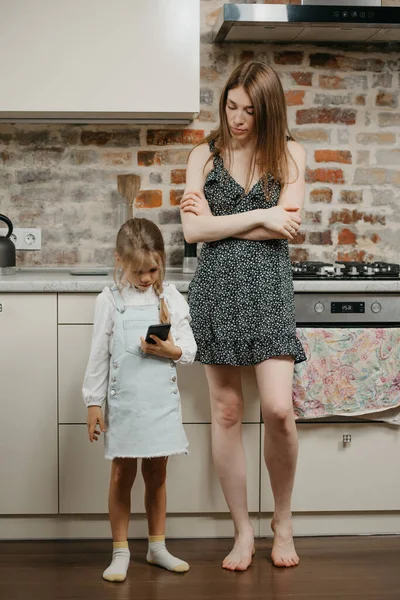 A blonde girl is gaming on a smartphone near her mother with pale skin in the kitchen. A caucasian mom is standing near a pretty daughter which is using a cellphone at home.