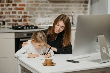 Genç, gülümseyen bir anne, iş yerindeki defterine çizim yaparken güzel bir kıza sarılıyor. Muhteşem bir anne, evde ödev yapan sarışın çocuğuna bakıyor..