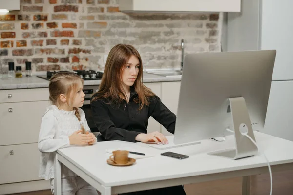 A mother is working remotely at home while a daughter is watching it. A businesswoman is working from an apartment near her blonde child.