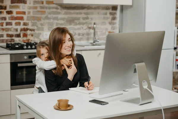 A young mother is working remotely at home while a daughter is hugging her. A smiling businesswoman is doing notes while working from an apartment near her blonde child.