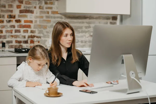 A young mom is working remotely carefully while a daughter is drawing in the notebook at home. A pretty mother is studying on a computer near her blonde child which is writing letters diligently.