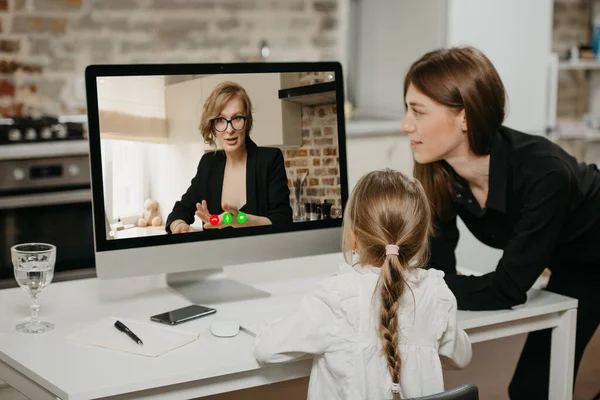 一位年轻的母亲和女儿在与一位女教师在线授课时 通过台式计算机上的视频通话 一位母亲看着她的金发孩子在家里远程学习 — 图库照片