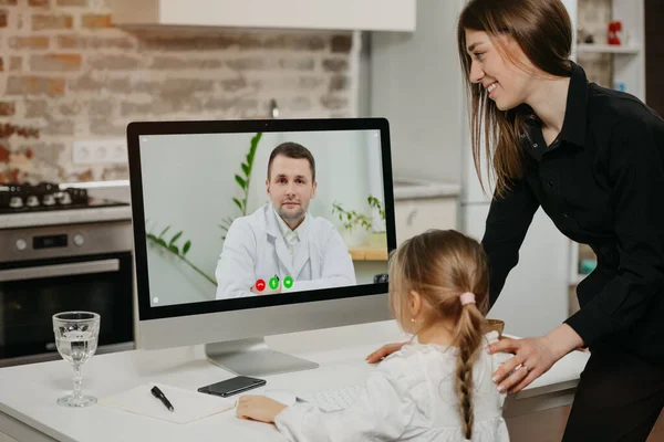 一位年轻的母亲和女儿在网上医生的预约过程中 通过台式计算机上的视频通话 一个母亲和一个金发碧眼的孩子在家里得到医生的远程咨询 — 图库照片
