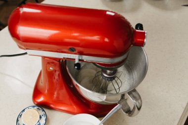 Mutfaktaki diğer malzemelerin yanında paslanmaz çelik bir çorba kasesinde yumurta beyazı kırbaçlayan kırmızı stand mikserinin yakın bir fotoğrafı.