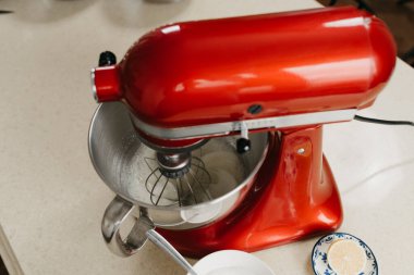 Mutfaktaki diğer malzemelerin yanında paslanmaz çelik bir çorba kasesinde yumurta beyazı kırbaçlayan kırmızı stand mikserinin yakın bir fotoğrafı.