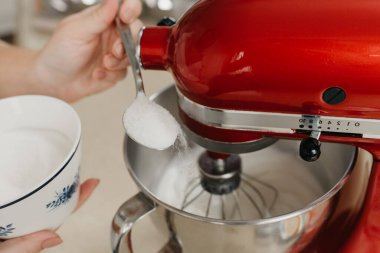 Kırmızı stand mikseri onu çırparken paslanmaz çelik çorba kasesine şeker kaşıklayan bir kadının yakın bir fotoğrafı.