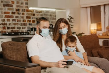 Baba, anne ve oğul, Coronavirus 'un (COVID-19) yayılmasını önlemek için tıbbi maske takmış bir kanepede oturuyorlar. Evde karantina altında bir aile. Akrabalar akıllı telefondaki içeriğin keyfini çıkarıyorlar