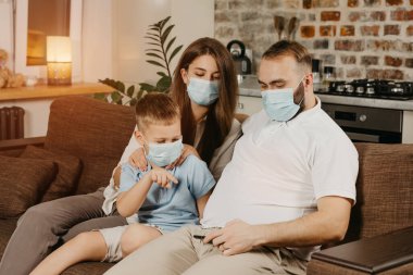 Baba, anne ve oğul, Coronavirus 'un (COVID-19) yayılmasını önlemek için tıbbi maske takmış bir kanepede oturuyorlar. Evde karantina altında bir aile. Akrabalar koltukta cep telefonuyla video izliyorlar.