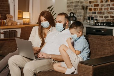 Baba, anne ve oğul, koronavirüsün (COVID-19) yayılmasını önlemek için yüz maskeleriyle kanepede oturuyorlar. Evdeki karantina sırasındaki akrabalar. Babam ailesine yakın bir laptopta çalışıyor..