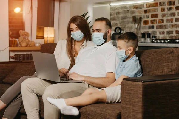 Dad, mom and son are sitting on the sofa in face masks to avoid the spread of coronavirus (COVID-19). Relatives during quarantine at home. Father is working remotely on a laptop near his family.