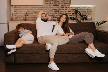 Baba ve anne kanepede televizyon seyrediyorlar. Oğul ise akşamları yakınlarda uyuyor. Aile evde..