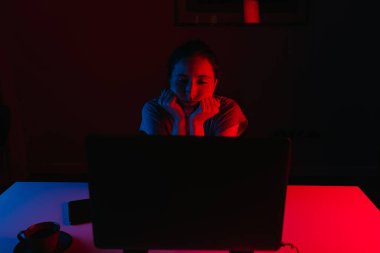 Evde uzaktan kumandalı bir bilgisayarla çalışan bir kadın var. Bir kız başını masaya yaslanmış ellerinin üzerinde dinleniyor. Bir bayan çalışan bilgisayarın önünde mavi ve kırmızı ışıkta sıkılıyor..