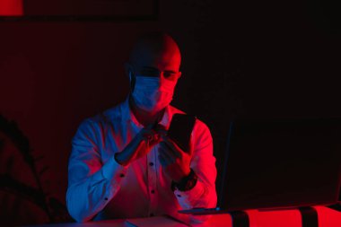 Tıbbi maske takan kel bir adam evde dizüstü bilgisayarla uzaktan çalışıyor. Kulaklıklı bir adam akıllı telefondan haberleri okuyor. Mavi ve kırmızı ışıkta bilgisayarın önünde çalışan bir erkek..