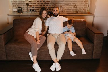 Baba, anne ve oğul akşamları kanepede televizyon seyrediyorlar. Memleketteki akrabalar. Oğlu, ailesinin yanında cep telefonuyla oyun oynuyor..