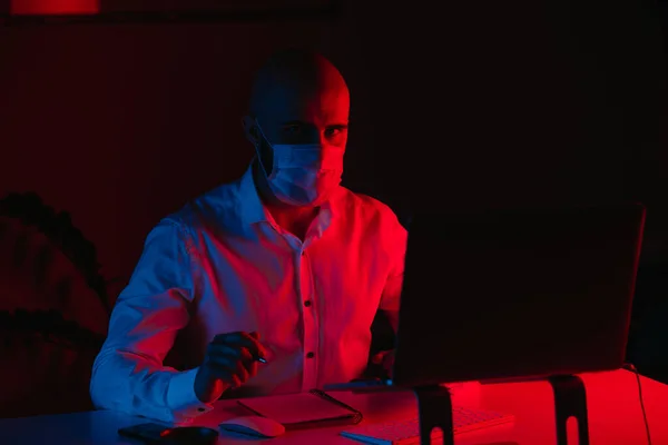 A bald man in a medical face mask is working remotely on a laptop at home. A guy is staring while doing notes. A male employee with a pen in front of the computer under blue and red light.