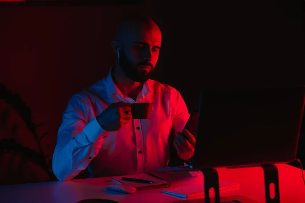 A bald man with a beard is working remotely on a laptop at home. A guy with earphones is drinking coffee and scrolling the news. An employee in front of the computer in the rays of blue and red light.