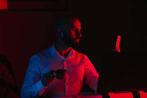 A man with a beard is working remotely on a laptop at home. A guy with earphones is reading the news on a smartphone. An employee with a cup in front of the computer in the rays of blue and red light.
