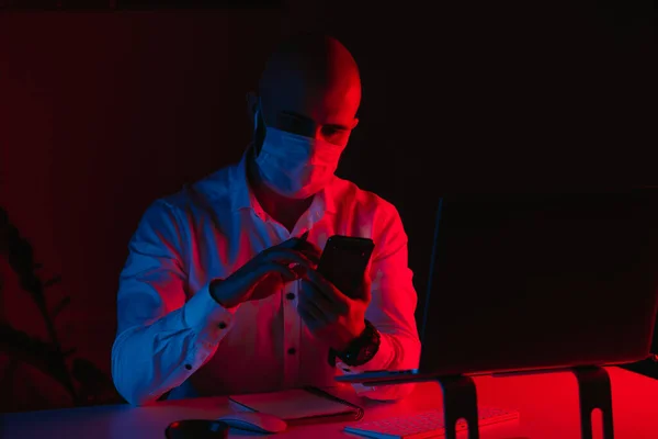 A bald man in a medical face mask is working remotely on a laptop at home. A guy is reading the news on a smartphone. A male employee with a pen in front of the computer under blue and red light.