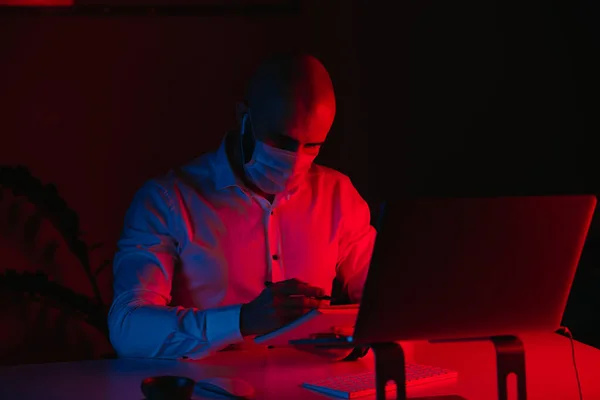 A bald man in a medical face mask is working remotely on a laptop at home. A guy with earphones is doing notes. A male employee with a pen in front of the computer under blue and red light.