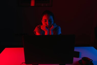 Evde uzaktan kumandalı bir bilgisayarla çalışan bir kadın var. Bir kız başını masaya yaslanmış ellerinin üzerinde dinleniyor. Bir bayan çalışan bilgisayarın önünde mavi ve kırmızı ışıkta sıkılıyor..