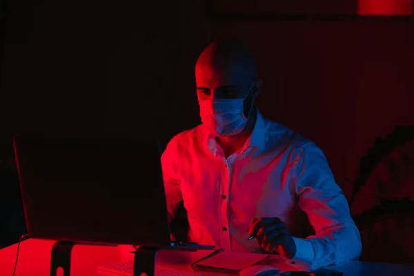A bald man in a medical face mask is working remotely on a laptop at home. A guy is reading and doing notes. A male employee with a pen in front of the computer under blue and red light.
