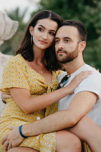 A close photo of a brunette girl in a yellow dress who is sitting on the legs of her boyfriend and hugging him on the ancient bridge in old Spain town. A couple of tourists on a date in Valencia.