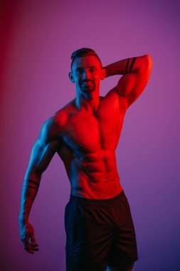 Sakallı kaslı bir adam mavi ve kırmızı ışığın altında koluyla poz veriyor. Atletik bir adam spor gövdesini gösteriyor. Antrenmandan sonra şort giyen bir sporcu..