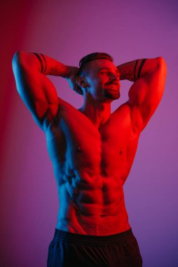 Sakallı kaslı bir adam mavi ve kırmızı ışığın altında kolları başının arkasında rahatlıyor. Atletik bir adam spor gövdesini gösteriyor. Antrenmandan sonra şort giymiş gülümseyen bir sporcu..