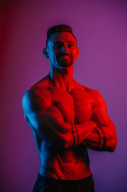 Sakallı kaslı bir adam mavi ve kırmızı ışığın altında katlanmış kollarıyla poz veriyor. Atletik bir adam spor gövdesini gösteriyor. Antrenmandan sonra şort giyen bir sporcu..