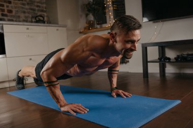 Kaslı bir adam evinde mavi yoga minderinin üzerinde şınav çekiyor. Kollarında dövme olan atletik bir adam evde göğüs ve triseps egzersizi yapıyor. Çıplak gövdeli bir vücut geliştirici.