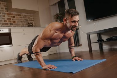 Kaslı bir adam evinde mavi yoga minderinin üzerinde şınav çekiyor. Kollarında dövme olan atletik bir adam evde göğüs ve triseps egzersizi yapıyor. Çıplak gövdeli mutlu bir vücut geliştirici.