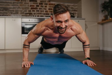 Sakallı, gülümseyen kaslı bir adam akşam dairesinde mavi yoga minderinde şınav çekiyor. Kollarında dövme olan atletik bir adam evinde göğüs ve triseps egzersizi yapıyor..