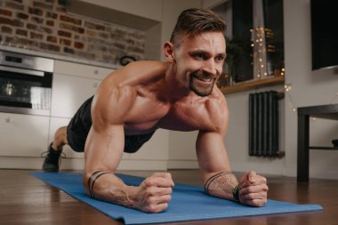 Çıplak gövdeli mutlu kaslı bir adam akşamları dairesinde mavi yoga minderinin üzerinde kalas yapıyor. Kollarında dövme olan gülümseyen atletik bir adam evde antrenman yapıyor..