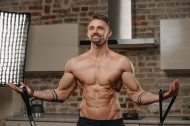 Kaslı bir adam dairesinde elastik bir çekme halatıyla çalışıyor. Kollarında dövmeleri olan atletik bir adam evde sportif fiziğini gösteriyor. Çıplak gövdeli bir sporcu.