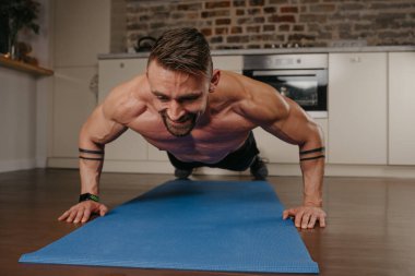 Sakallı mutlu kaslı bir adam akşamları evinde mavi yoga minderinde şınav çekiyor. Kollarında dövme olan atletik bir adam evinde göğüs ve triseps egzersizi yapıyor..