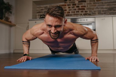 Kaslı bir adam akşamları evinde mavi yoga minderinin üzerinde şınav çekiyor. Kollarında dövme olan atletik bir adam evde göğüs egzersizi yapıyor. Çıplak gövdeli bir vücut geliştirici.