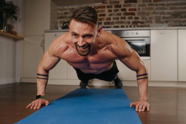 Sakallı, gülümseyen kaslı bir adam akşam dairesinde mavi yoga minderinde şınav çekiyor. Kollarında dövme olan atletik bir adam evinde göğüs ve triseps egzersizi yapıyor..