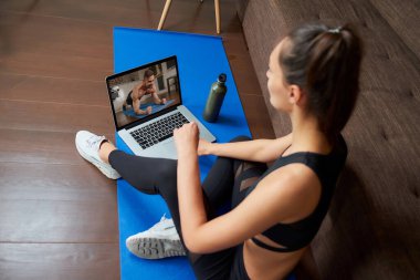 Bir kadının omzunda dizüstü bilgisayar görüntüsü. Dar takım elbiseli sporcu bir kız evinde dizüstü bilgisayardan egzersiz videosu izliyor. Kadın evde sanal fitness dersi sırasında koçu dinliyor.
