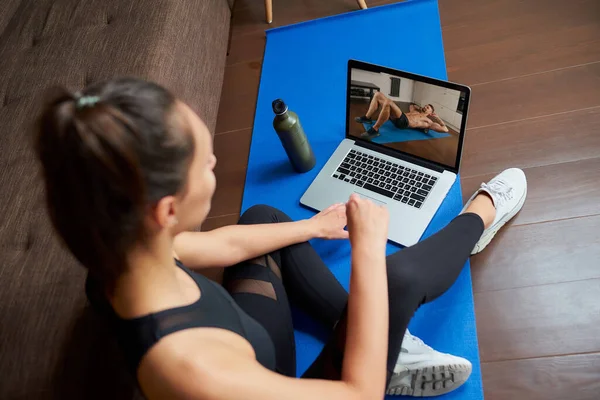 Bir kadının omzunda dizüstü bilgisayar görüntüsü. Dar takım elbiseli fit bir kız evinde dizüstü bilgisayarından egzersiz videosu izliyor. Kadın evdeki bir spor salonunda koçu dinliyor.