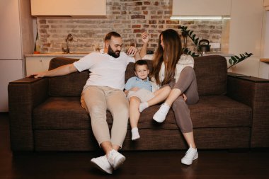 Baba, oğul ve anne evdeki kanepede sıkıcı TV programları izliyorlar. Ebeveynler akşamları oğullarına bakıyor. Memleketteki akrabalar. Uzun saçlı genç bir anne gülümsüyor..